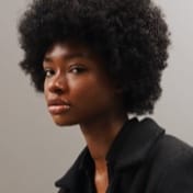Jessica Randall with natural curly hair wearing a dark jacket, sitting upright against a plain light gray background, neutral and calm demeanor